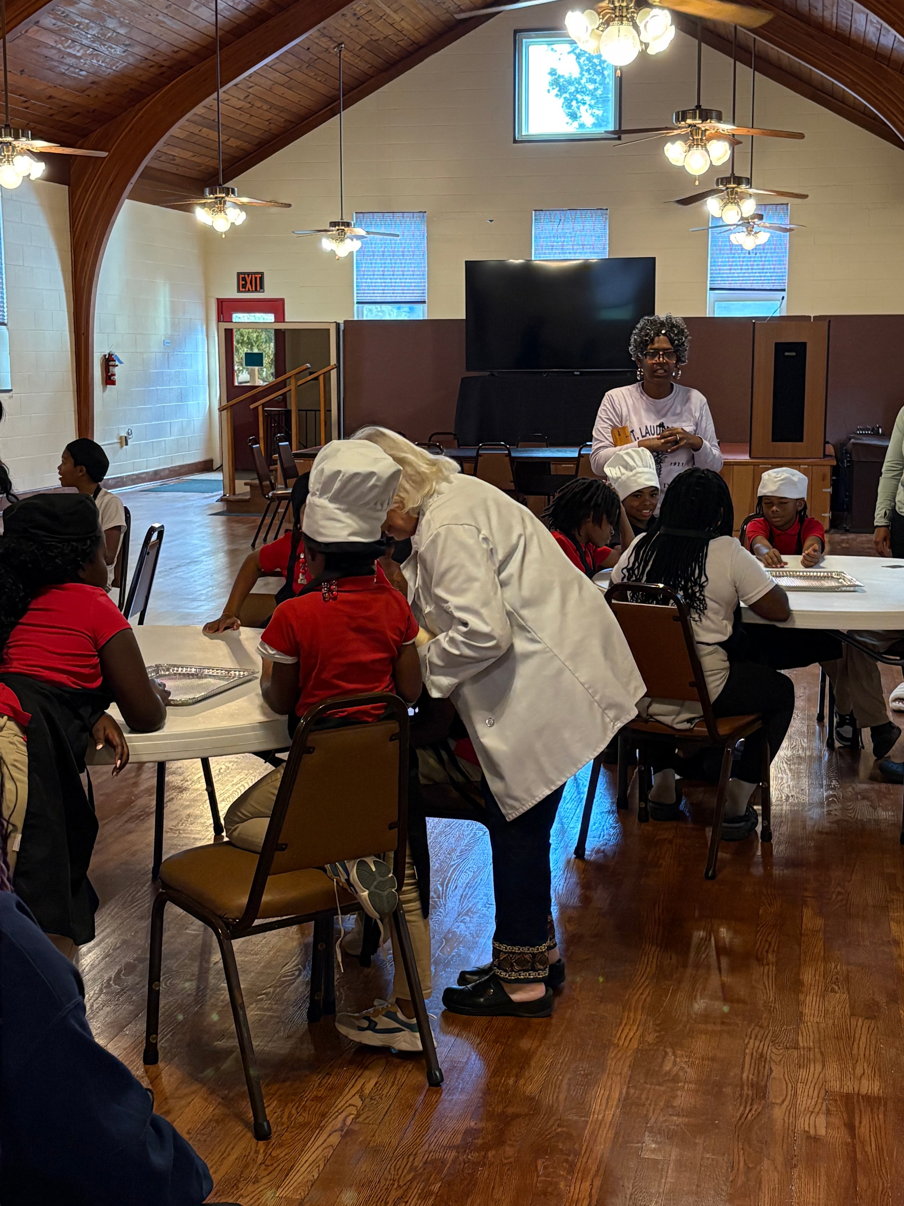 Young chefs in aprons and chef hats gathered around tables in the parish hall during a Kids R Cookin' session