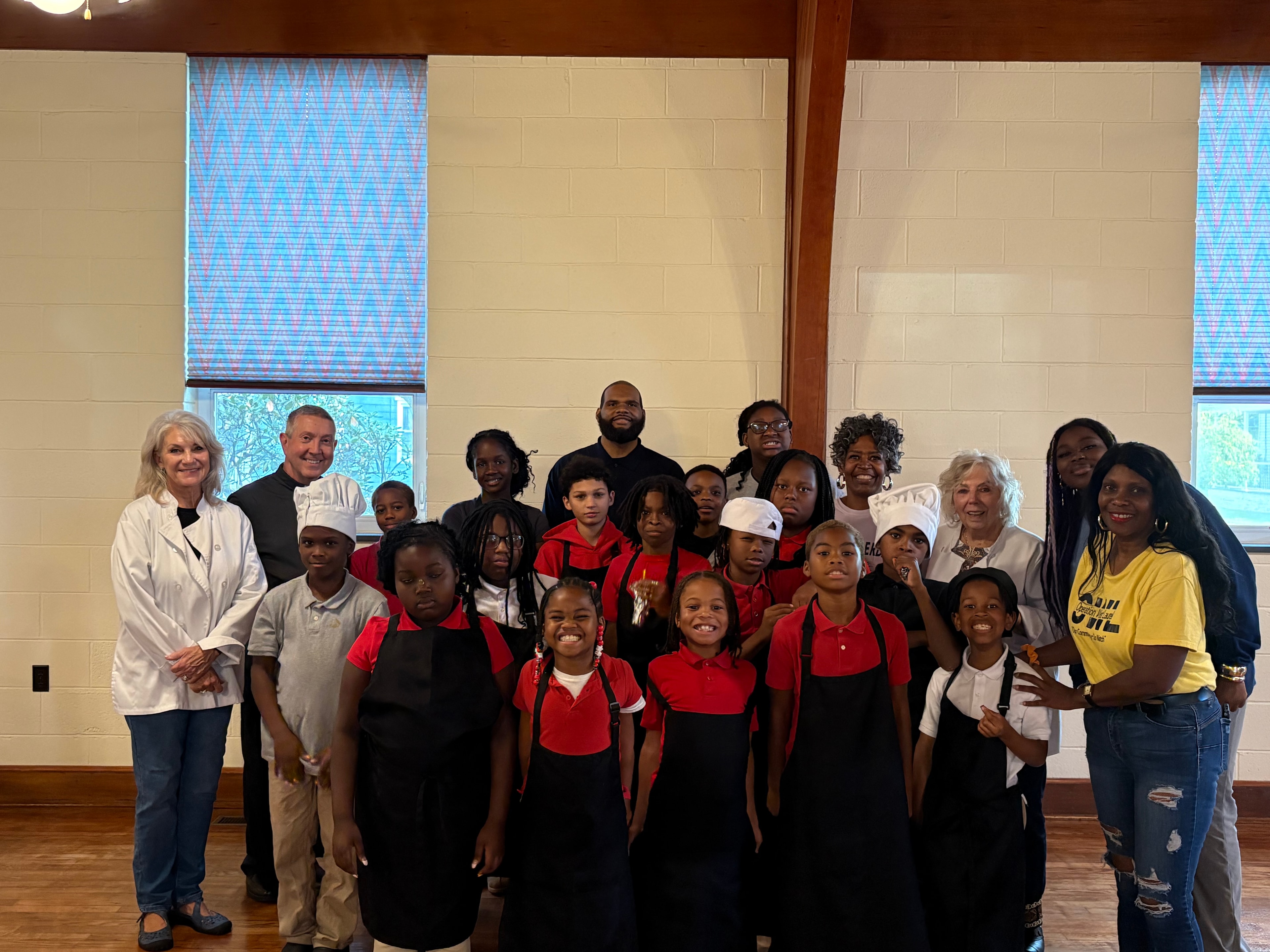 A group photo of Kids R Cookin' young chefs, volunteers, and leaders in aprons and chef hats in the parish hall