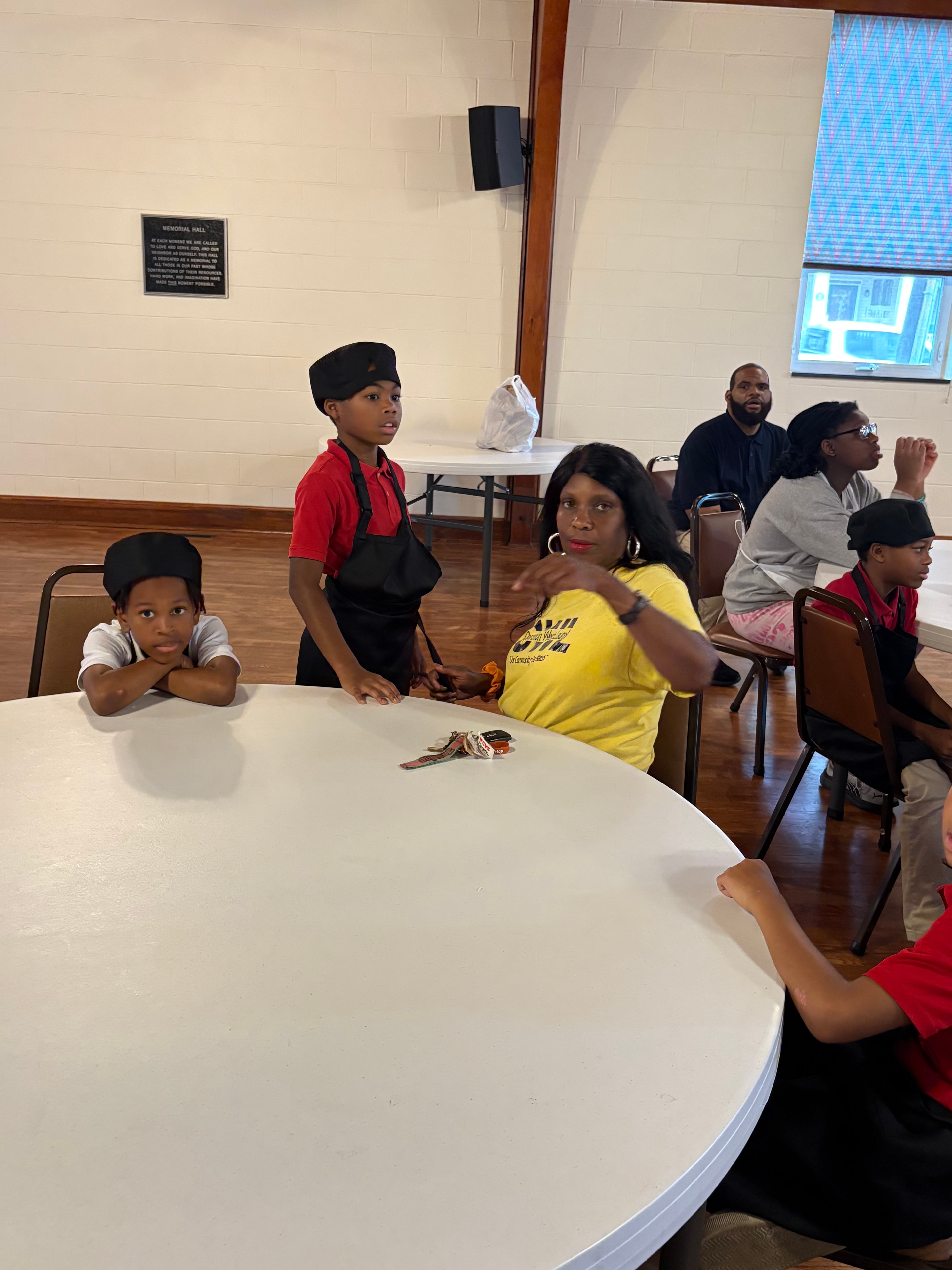 A volunteer leading a Kids R Cookin' lesson with children gathered around a table in the parish hall